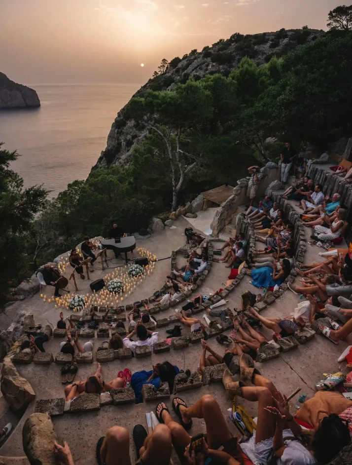 Público sentado en gradas de piedra frente al mar durante un concierto al atardecer